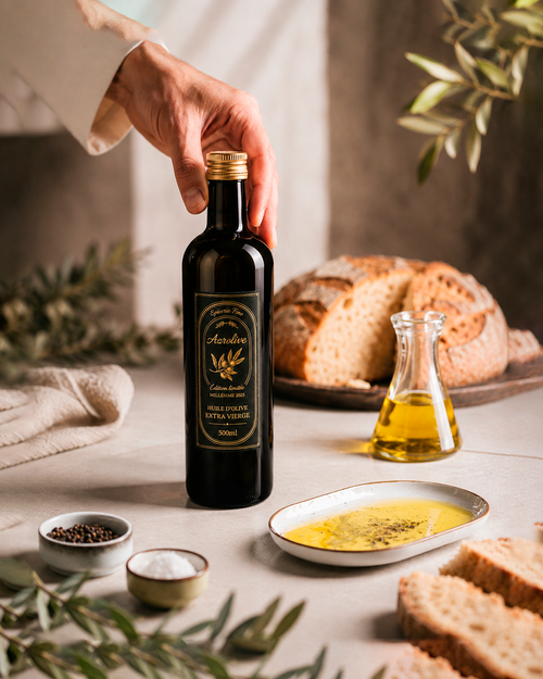 La bouteille Azrolive 500ml édition limitée saisie d'une main assurée, entourée de pain artisanal et d'olivier. Une scène qui allie élégance et authenticité kabyle.