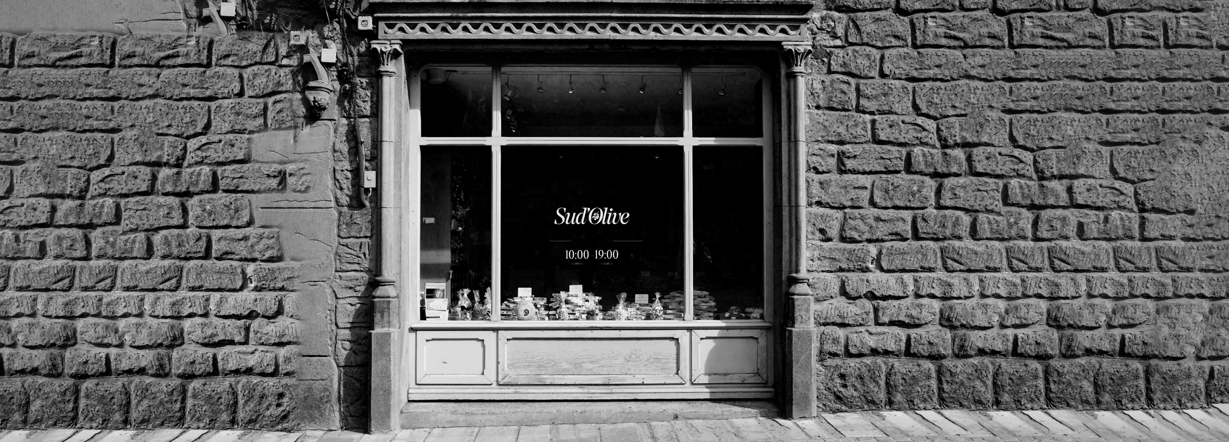 Façade en pierre de la boutique Sud'Olive, épicerie fine spécialisée en huile d'olive kabyle, avec vitrine élégante et horaires affichés, photographie noir et blanc